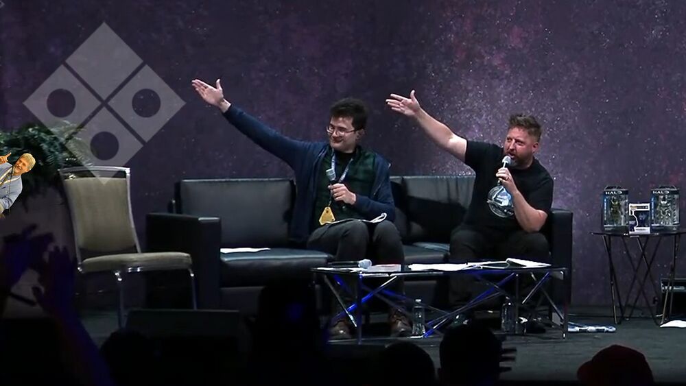 Image of Alex Wakeford and Jeff Easterling on the Community Stage at the Halo World Championship 2023 gesturing towards a seat with the Canon Fodder logo and a mini Troy Denning also pointing at it